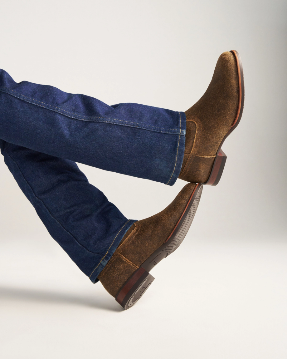 The Glen in Dustwood Roughout. Person wearing blue jeans and Dustwood Roughout boots with crossed legs, photographed against a plain light background.