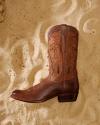 A single brown leather cowboy boot with decorative stitching lies on sandy ground.