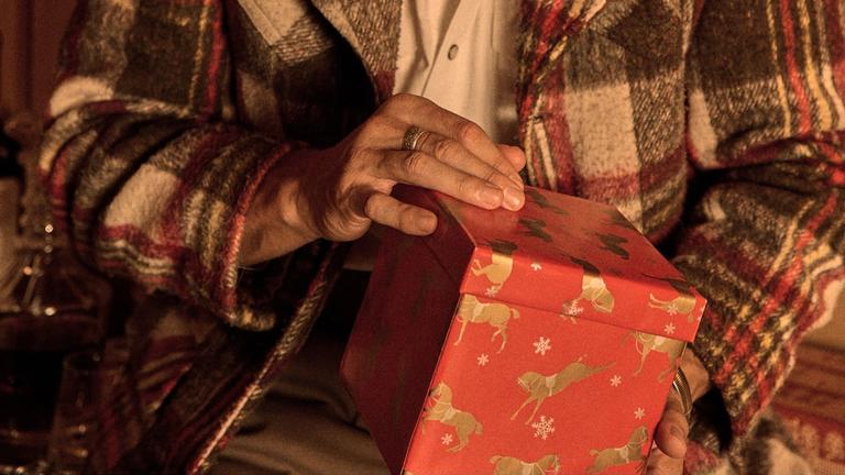 A person wearing a plaid jacket holds a red gift box decorated with images of reindeer and snowflakes.
