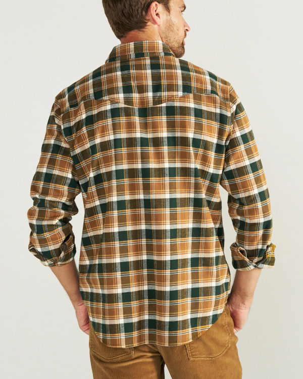 A man stands facing away, wearing a brown, green, and white plaid long-sleeve shirt with brown pants against a plain light background.