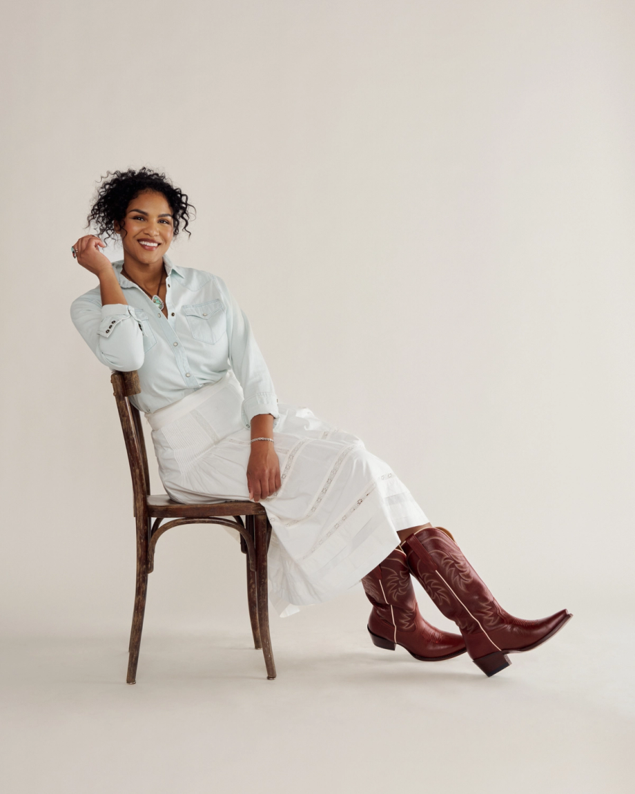 A woman sits sideways on a wooden chair, smiling, wearing a light blue shirt, white skirt, and brown cowboy boots against a plain background.