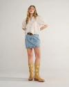 A woman stands against a plain background wearing a white blouse, denim skirt, brown belt, and tan cowboy boots with white circular patterns.