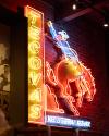 Neon sign on a brick wall displays the word “Tecovas” vertically and depicts a cowboy riding a bucking horse with the words “Western Wear” underneath.