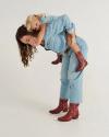 An adult bends forward giving a piggyback ride to a child. Both wear light blue denim outfits and red boots. The background is plain white.