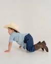 A young child wearing a cowboy hat, plaid shirt, jeans, and cowboy boots crawls on hands and knees against a plain white background.