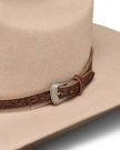 Close-up of a beige cowboy hat featuring a brown leather band with tooled patterns and a decorative silver buckle.