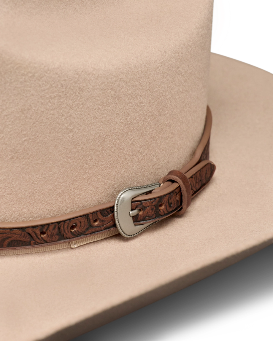 Close-up of a beige cowboy hat featuring a brown leather band with tooled patterns and a decorative silver buckle.