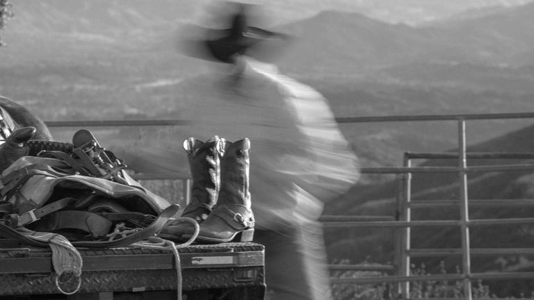 Black and white image of a blurred figure in a cowboy hat standing near a fence, with boots and riding gear on a truck bed in the foreground.