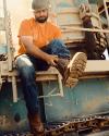 A man wearing an orange shirt, jeans, and a hard hat sits on industrial equipment, pulling on a cowboy boot.