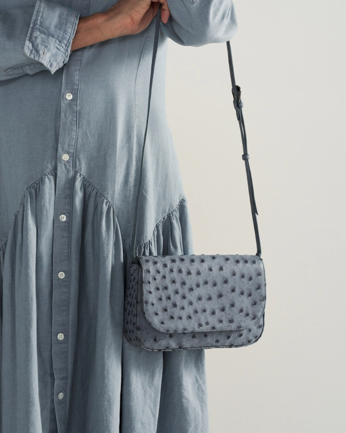 A person wearing a light blue buttoned dress holds a textured blue shoulder bag with an adjustable strap.