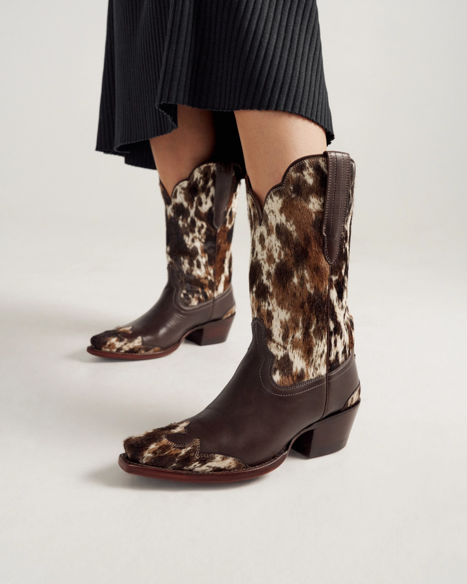 Person wearing a black skirt and The Sadie Hair on Hide / Cowhide-patterned cowboy boots with pointed toes and chunky heels, standing on a plain white background.
