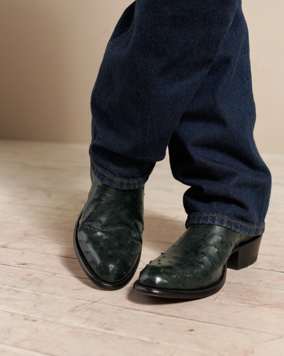 Person wearing dark green leather boots and blue jeans standing on a light wooden floor.