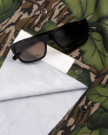 Black rectangular sunglasses rest on a camouflage-patterned shirt with green leaf and brown stripe designs, partially folded to reveal a white interior and metal snap buttons.