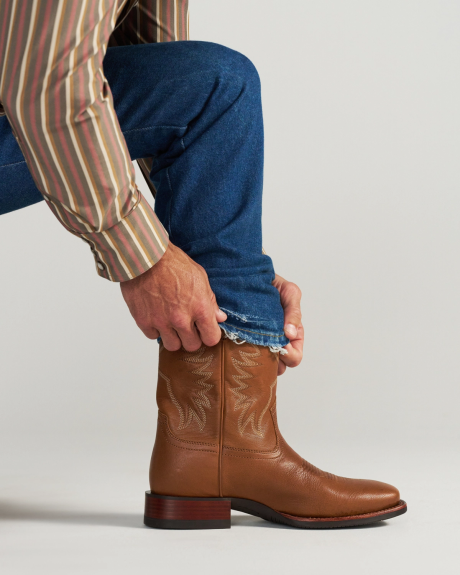 The Dusty in Mesa Goat. Person wearing a striped shirt and blue jeans, pulling a pant leg over a tan leather cowboy boot with decorative stitching, ready to explore the rugged trails of Mesa.