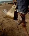 Close-up of a person wearing worn leather boots and jeans, foot resting in a metal stirrup, with a dusty outdoor setting in the background.