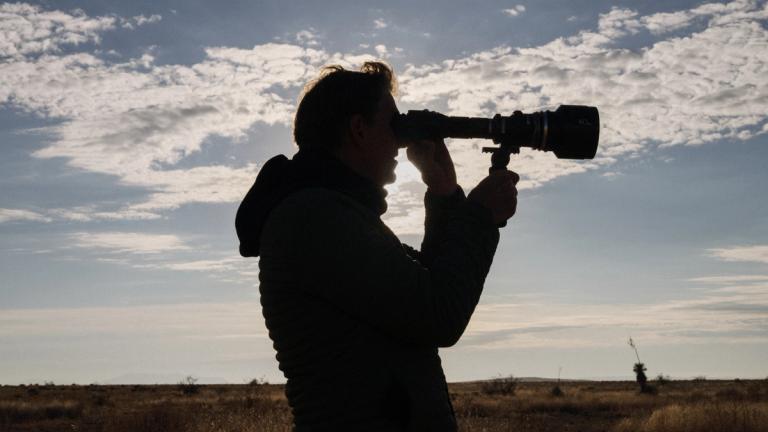 A person stands outdoors in a field, silhouetted against the sky, holding a camera with a large telephoto lens and looking through the viewfinder.