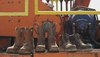 Four pairs of worn boots lined up on an industrial orange platform, with machinery visible in the background.