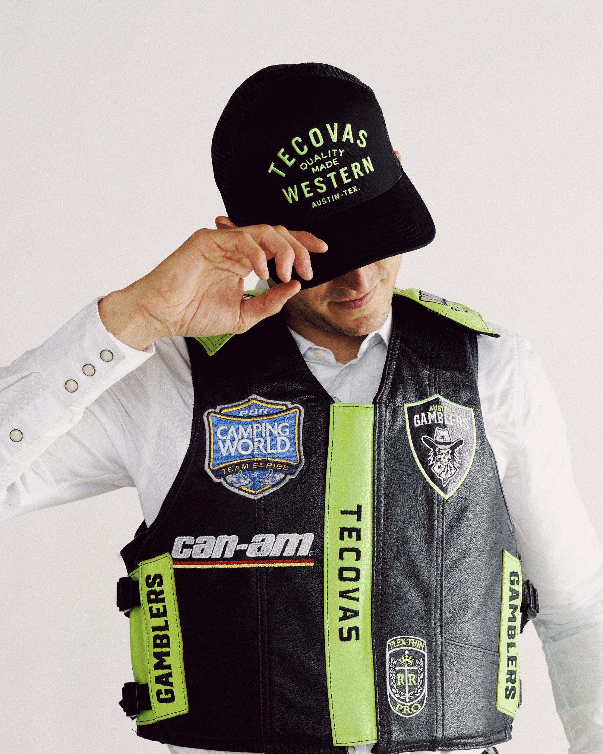 A person in a white shirt wearing a black and green branded vest and a matching Tecovas cap, with their hand holding the brim of the cap.