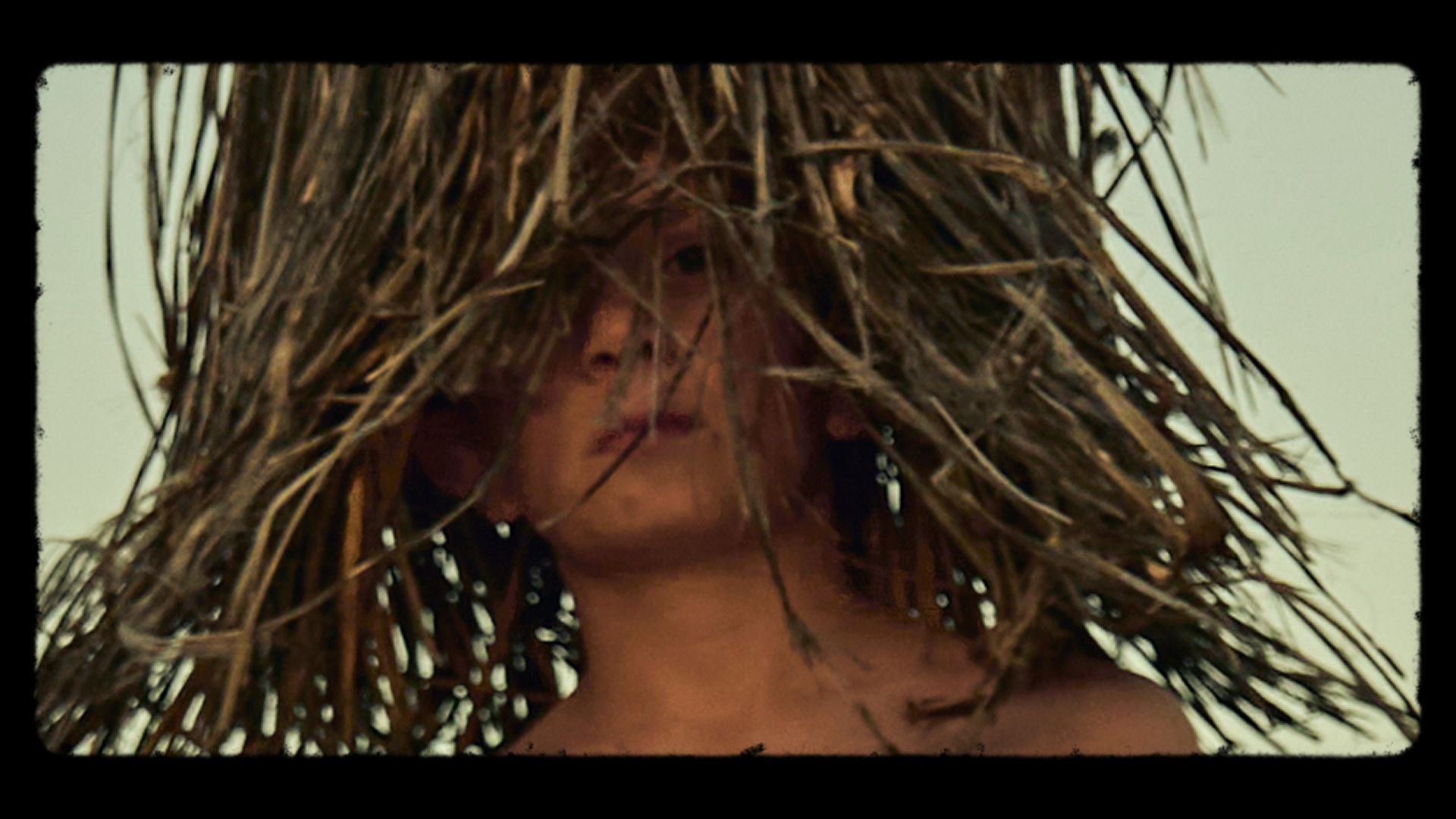 A child with a neutral expression looks through hanging dry grass or straw, partially obscuring their face.