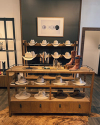 Wooden display shelves with white and beige cowboy hats, brown cowboy boots, and accessories in a retail store with a dark accent wall and framed art above.