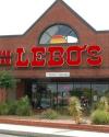 A brick storefront with large red letters spelling "LEBOS" above a glass entrance. Desert plants are in front, and a neon "OPEN" sign is visible in the window.