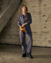 A person in a blue coverall and black women's work boots stands indoors by a concrete wall, holding a pair of work gloves and looking slightly to the side.
