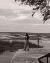 A person stands alone on a wooden dock, looking out over a marshland under a cloudy sky, with water and tall grasses stretching into the distance.