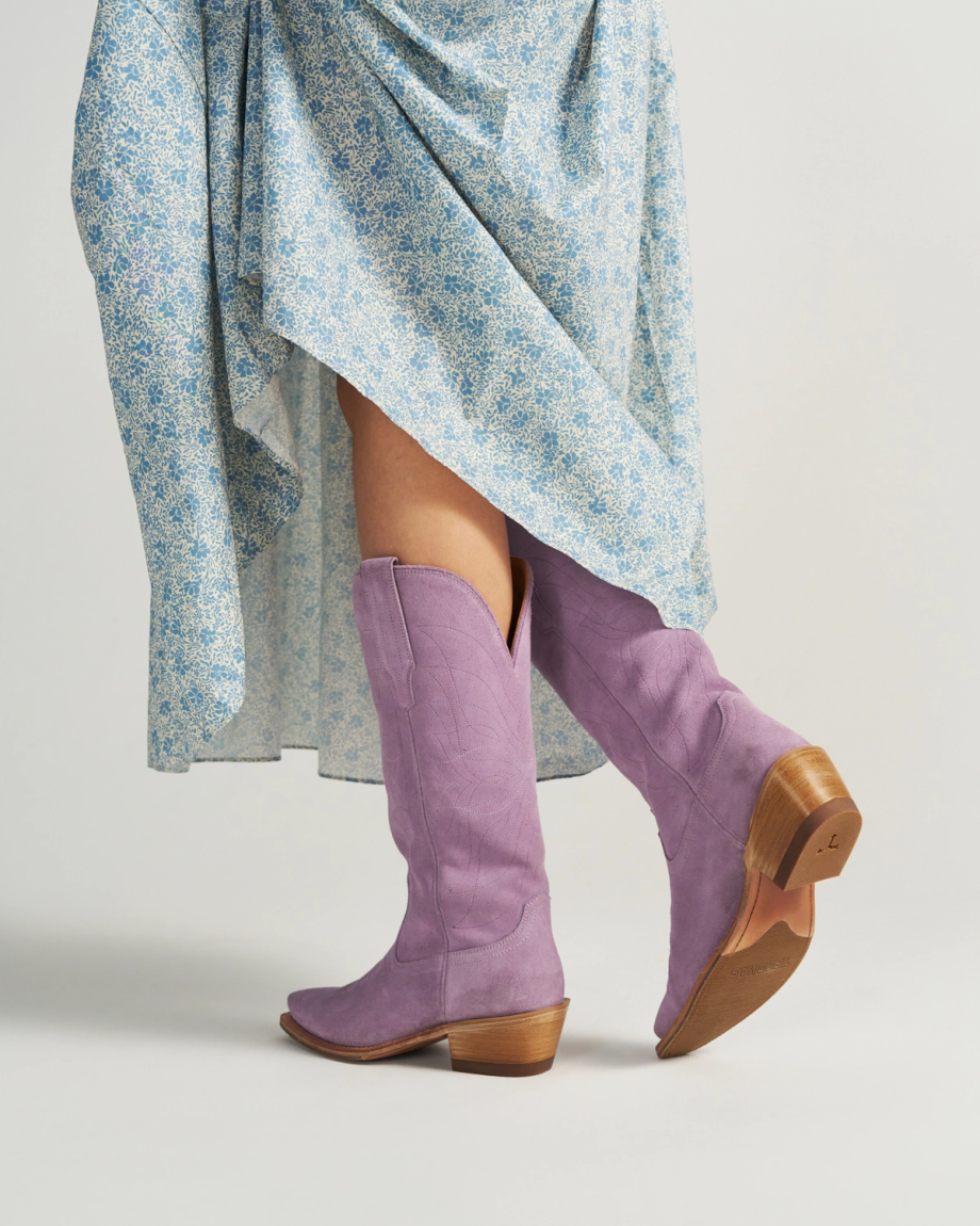 A person in a blue floral dress wears The Annie Suede knee-high lavender cowboy boots with wooden heels, posing against a plain background.