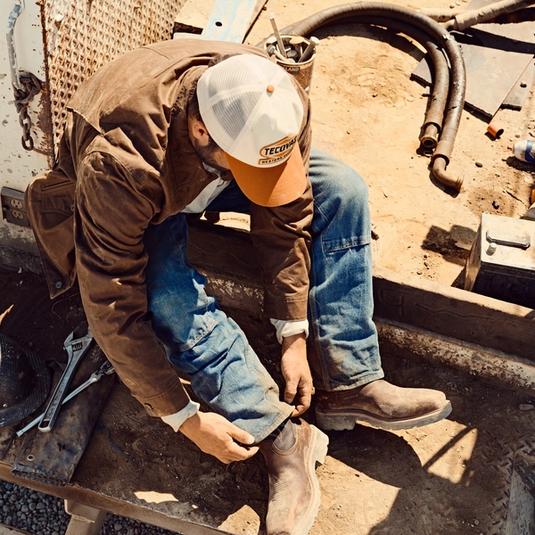 A person in work clothes and a cap sits outdoors, bending down to pull on a brown boot, with tools and equipment scattered around on a dusty surface.