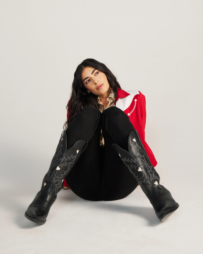 A woman with long dark hair sits on the floor, leaning back with her knees up, wearing a red jacket, black pants, and The Annie Cowhide Midnight cowboy boots against a plain white background.
