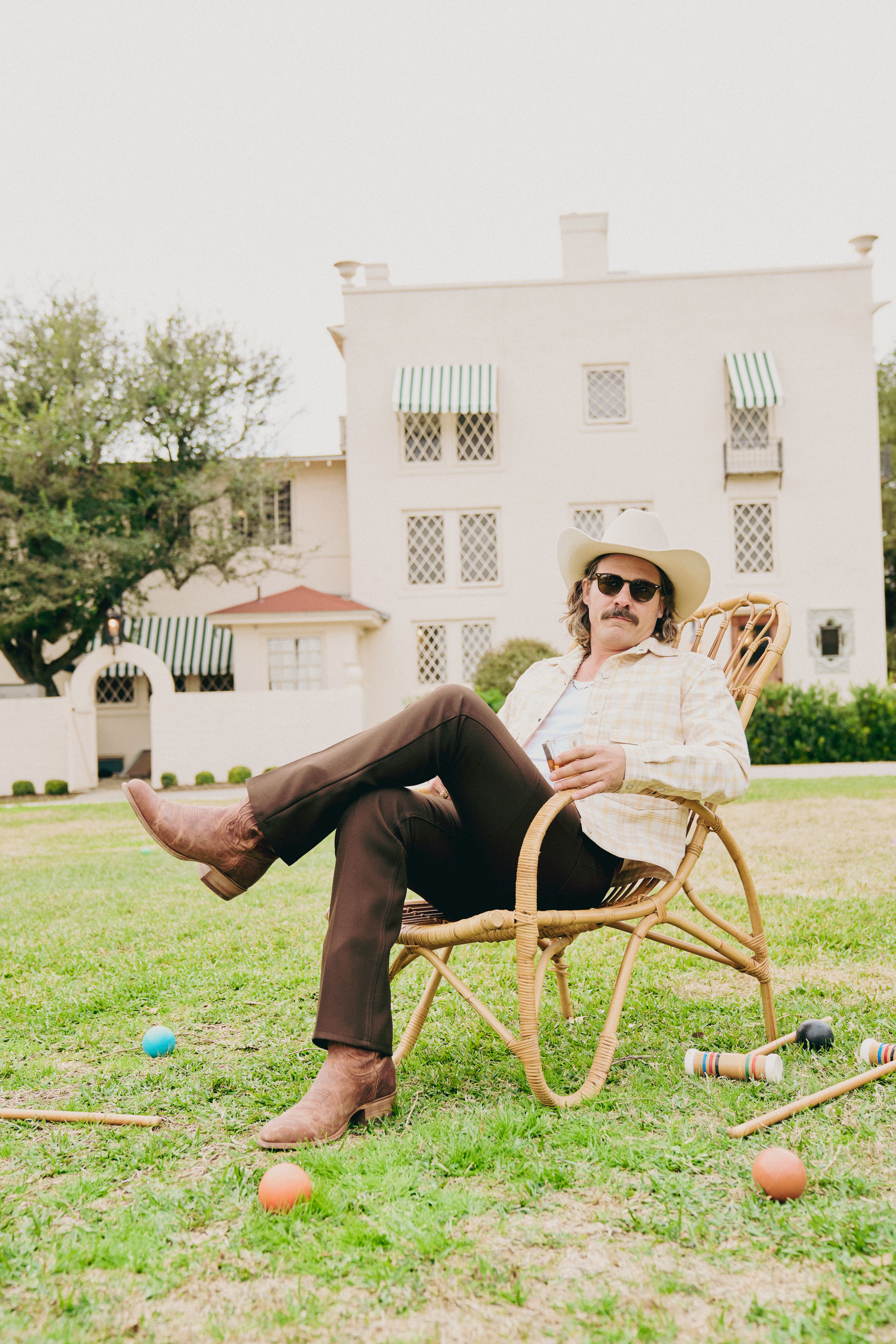A man wearing a cowboy hat, sunglasses, and boots sits in a wicker chair on a lawn, with croquet balls and mallets scattered nearby and a house in the background.