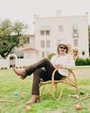 A man wearing a cowboy hat, sunglasses, and boots sits in a wicker chair on a lawn, with croquet balls and mallets scattered nearby and a house in the background.