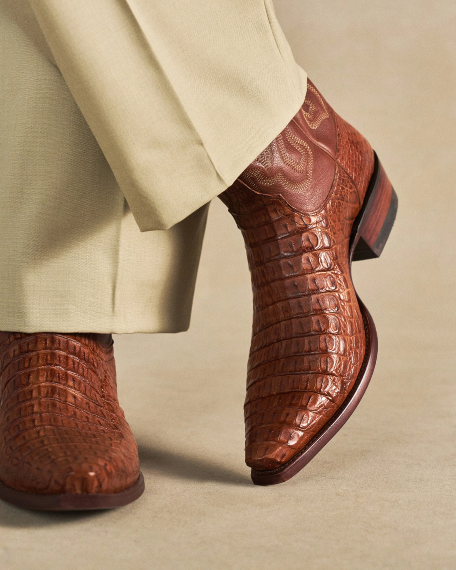 The Garrett in Pecan Hornback Caiman. A person wearing beige pants and brown textured cowboy boots, specifically The Garrett Hornback Caiman, stands on a light tan surface.