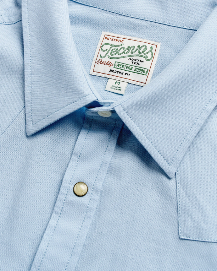 Close-up of a light blue collared shirt with a visible label inside the collar. The shirt features a button near the neckline.