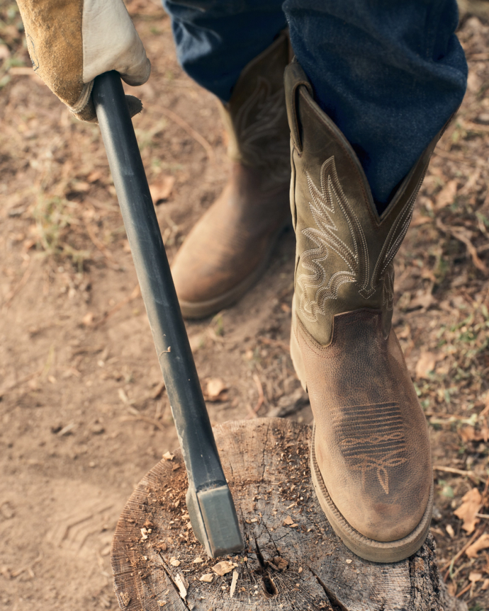 A person with boots—the Rugged Round Toe Cowhide in Umber / Sage—and a shovel.