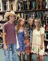 Four people wearing casual outfits and cowboy boots stand in front of shelves filled with various cowboy boots in a store.