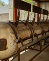 A row of large outdoor metal barbecue grills with lids, showing signs of rust and use, lined up under a covered area with a tree and building in the background.