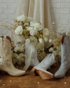 White and beige cowboy boots next to a wedding bouquet against a brick backdrop with white flowers on the floor.