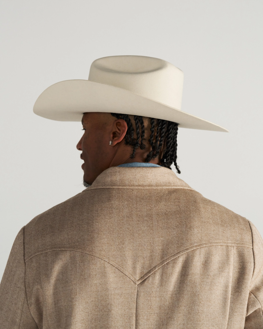 A person with braided hair, wearing a light-colored cowboy hat and a tan jacket, is shown from behind against a plain background.