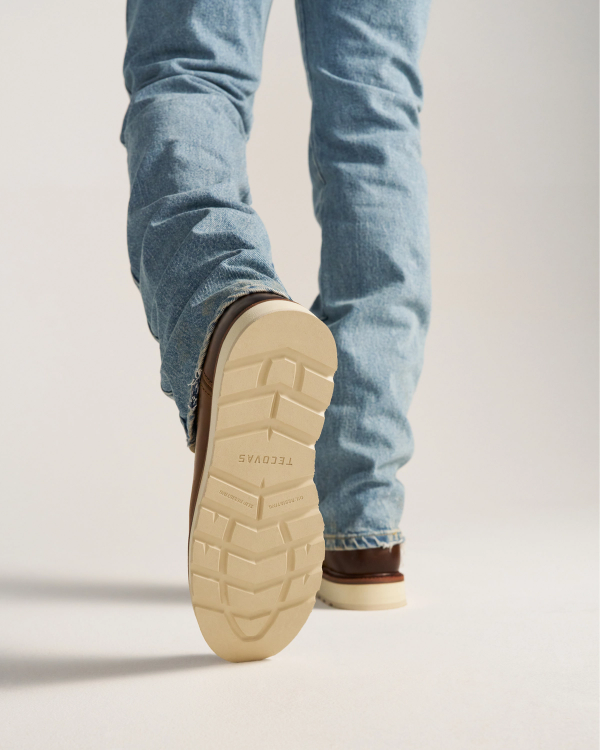 The Knox in Wheat Cowhide. Person wearing light blue jeans and brown shoes, shown from behind, with one foot lifted to display the sole marked with "TECOVAS," featuring the signature Wheat Cowhide finish.