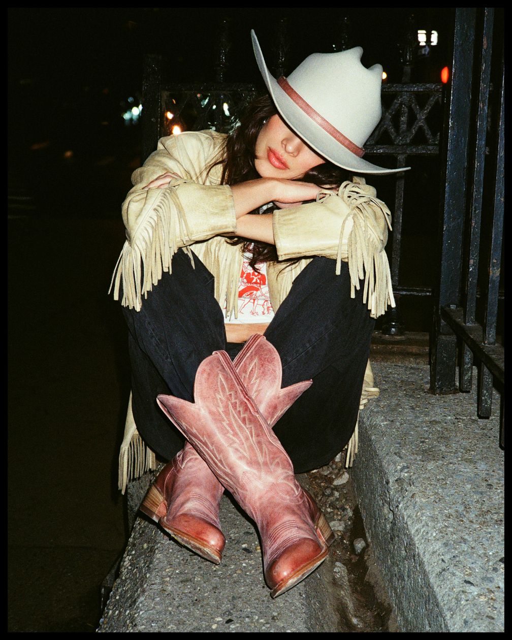A person in a white cowboy hat, fringe jacket, and pink cowboy boots sits on a curb at night with arms crossed on their knees.
