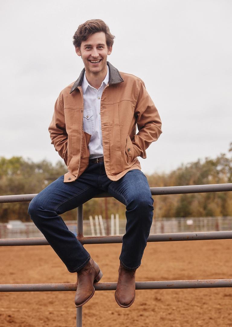 A young man sitting on a fence at a rodeo in a cowboy hat.
