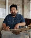 A man with a beard and mustache wearing a denim shirt works with brown materials at a table in an industrial setting with concrete block walls.