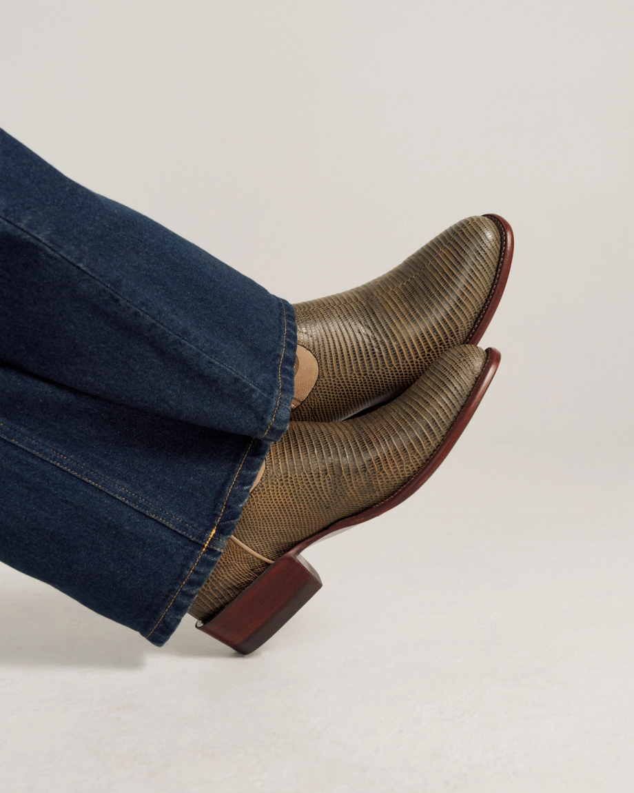 A person wearing blue denim jeans and textured brown ankle boots with a block heel sits with feet crossed against a plain background.