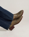 A person wearing blue denim jeans and textured brown ankle boots with a block heel sits with feet crossed against a plain background.