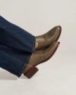 A person wearing blue denim jeans and textured brown ankle boots with a block heel sits with feet crossed against a plain background.