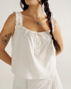 A woman wearing a white sleeveless blouse with scalloped edges and buttons, a silver necklace with a green pendant, and a long braid. Floral tattoos are visible on her arms.