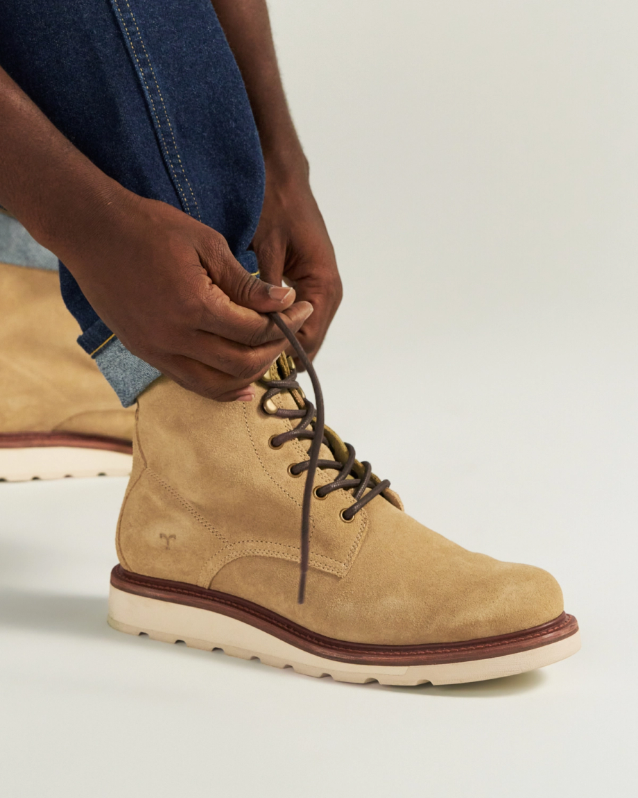 The Knox Lace Up Shearling in Sandstone Roughout. Person tying the brown laces of The Knox Lace Up Shearling Roughout tan suede boot with a white sole, wearing blue jeans, against a neutral background.