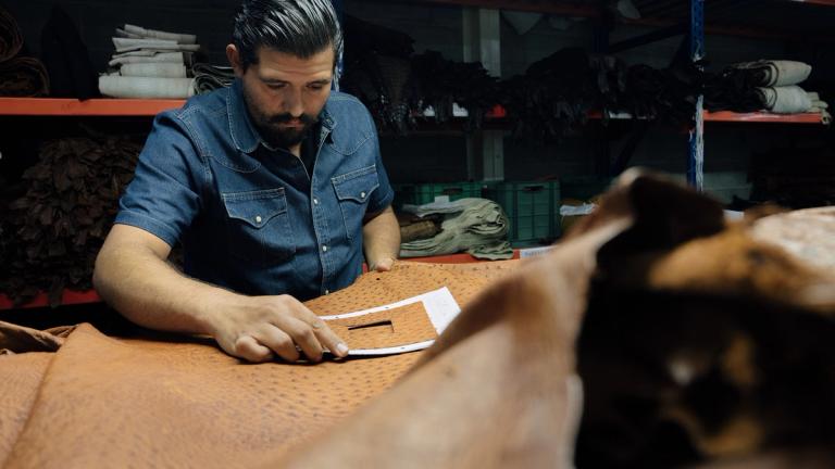 A man in a denim shirt measures a piece of brown leather with a template in a workshop lined with shelves of materials.
