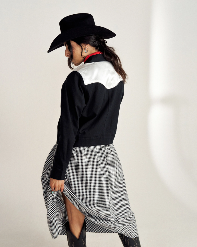 Person wearing a black cowboy hat, black and white jacket, and checkered skirt stands with their back turned.